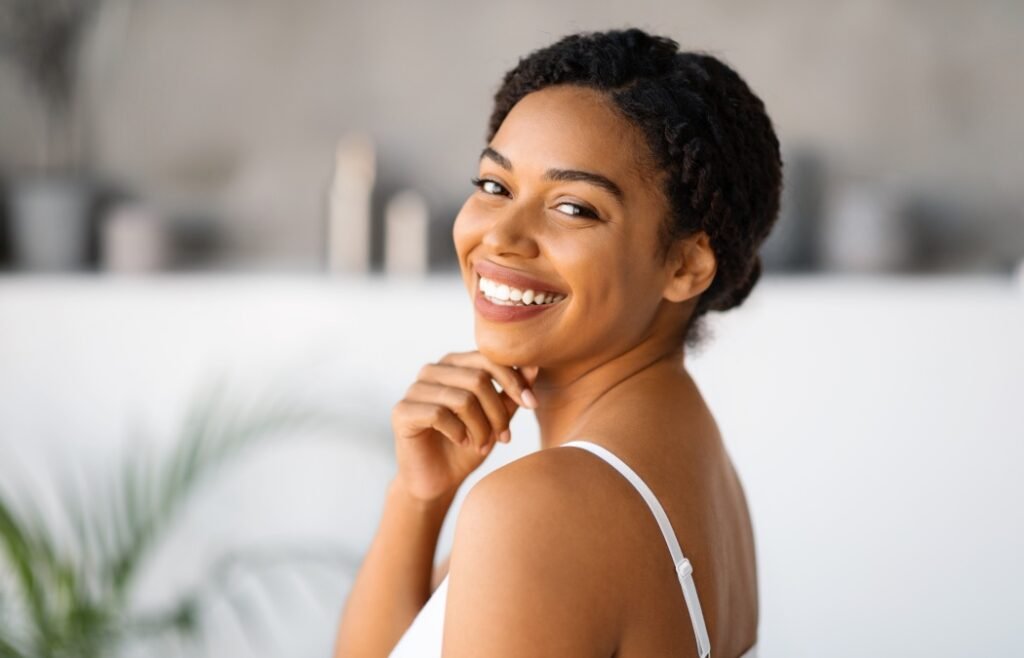 Natural Beauty. Attractive Black Woman Touching Chin And Looking At Camera While Standing In Bathroom At Home, Beautiful African American Female Enjoying Her Smooth Skin After Skincare Routine
